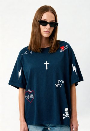 Femme aux cheveux bruns raides portant des lunettes de soleil noires et un t-shirt oversize foncé avec des symboles graphiques blancs et rouges incluant une croix, des cœurs et un crâne.