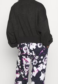 Černá oversized bomber bunda s texturovaným krokodýlím vzorem, spárovaná s multicolor květinovým vzorem tepláky na tmavém pozadí.