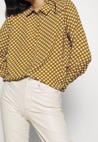 Blusa gialla senape con motivi floreali bianchi, colletto abbottonato e maniche lunghe, abbinata a pantaloni fitted color crema.