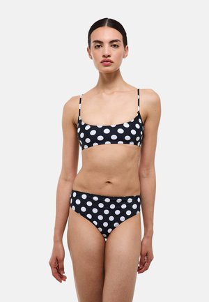 Femme portant un bikini noir et blanc à pois avec des bretelles fines, debout devant un fond gris clair uni.