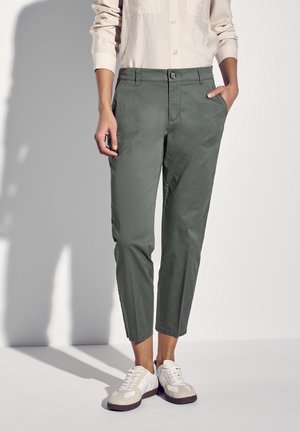 Persoon draagt olijfgroene cropped broek, beige overhemd met lange mouwen en knopen, en witte sneakers, met één hand in de zak.