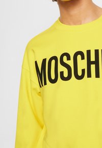 Osoba ubrana w jaskrawo żółtą bluzę z dużym, czarnym napisem „MOSCHINO” na klatce piersiowej, kadrowana tak, że widoczna jest tylko górna część tułowia.