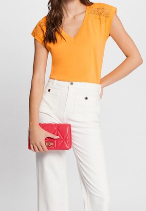 Frau trägt ein oranges V-Ausschnitt-Oberteil mit Spitzenärmeln, weiße hoch taillierte Hose, hält eine rote gesteppte Clutch.