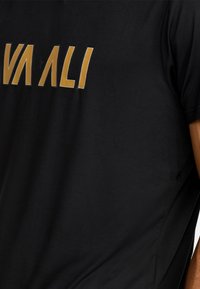 Zwarte t-shirt met grote, vetgedrukte gele letters die "VAALI" spellen. De stof lijkt zacht met een gladde textuur.