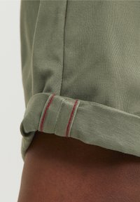 Olijfgroene katoenen shirt met opgerolde mouwmanchetten. De manchetten hebben een rode stikselafwerking. Soepele textuur en een casual ontwerp.