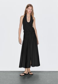 Zwarte halterjurk met een flowy, enkel-lange rok met een split aan de voorkant. Draagt zwarte slippers op een effen achtergrond.