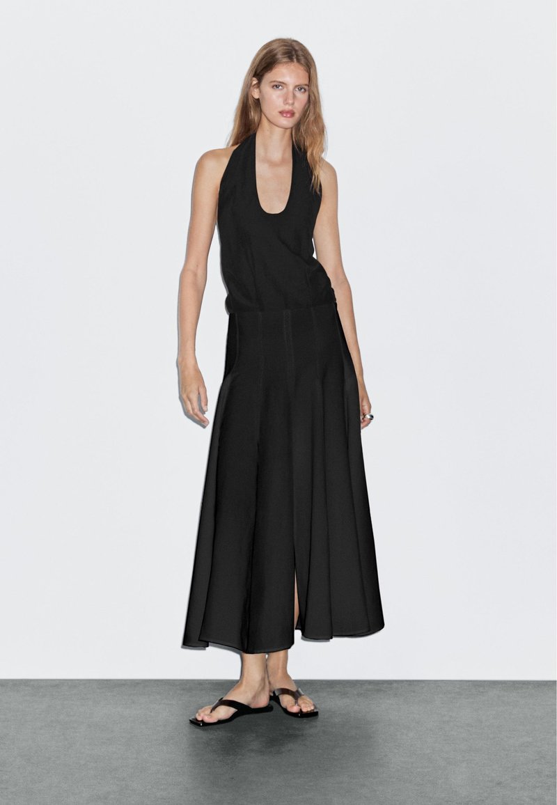 Sort halterneck-kjole med en flagrende, ankellang nederdel, der har en slids foran. Båret med sorte flip-flops på en ensfarvet baggrund.