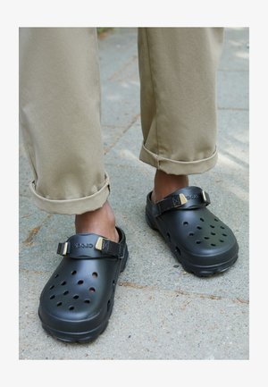 Black plastic clogs with ventilation holes, featuring a back strap and gold accents. Worn with rolled-up khaki pants on a textured surface.