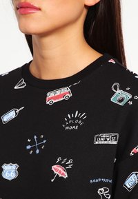 Sweatshirt noir avec des graphiques imprimés colorés, y compris une camionnette vintage, une boussole et divers icônes sur le thème du voyage, avec un col rond.