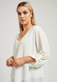 Vit chiffongblus med V-ringning och långa, uppvikta ärmar som hålls på plats med manschettknappar. Slät textur, lös passform och en diskret design.