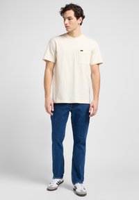 Krämfärgad bomullströja med bröstficka, kombinerad med blå denimjeans och vita skor. Enkel design, korta ärmar, avslappnad passform.