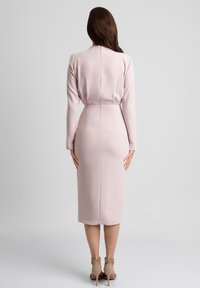 Różowa sukienka o średniej długości z długimi rękawami, dopasowanym kroju i gładkiej fakturze, z zapięciem na zamek z tyłu. Neutralne sandały na obcasie.