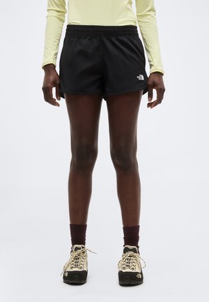 Person trägt schwarze North Face Shorts, beige Wanderschuhe, dunkle Socken und ein hellgelbes langärmliges Oberteil und steht vor einem weißen Hintergrund.