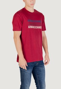 T-shirt in cotone bordeaux a maniche corte. Presenta il testo "ARMANIEXCHANGE" in colori a contrasto, disposto in un design sovrapposto, sul davanti.