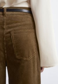 Brown corduroy pants with vertical ribbing, featuring two back pockets and a black belt, paired with a cream-colored blouse.