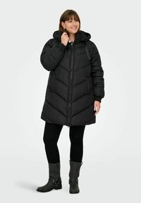 Black puffer coat with a hood, featuring a chevron design, front zipper, and ribbed cuffs. Paired with black trousers and ankle boots.