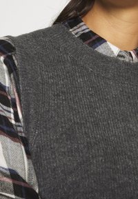 Grijs gebreid truif met een ribbelstructuur, gecombineerd met een geruite overhemd met rode, blauwe en zwarte strepen onder de kraag.