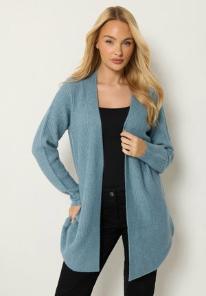 Blonde vrouw die een lange blauwe gebreide cardigan draagt over een zwarte top en zwarte broek, staand met één hand in de zak en één hand bij de borst.
