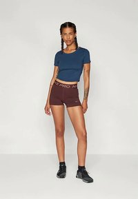 Haut court bleu marine associé à des shorts Nike Pro bordeaux. Chaussures de sport noires. Le modèle a les cheveux tressés et des tatouages visibles sur les bras.
