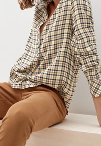 Camicia a scacchi nei toni giallo, nero e marrone, con vestibilità rilassata, design con bottoni e maniche arrotolate. Pantaloni marroni visibili sotto.