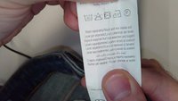 Bijela oznaka za njegu s uputama i simbolima na više jezika, uključujući smjernice za pranje i peglanje. Oznaku drži ruka.