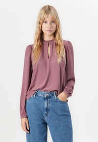 Donna bionda con capelli sciolti indossa una blusa mauve a maniche lunghe con scollatura a chiave e jeans blu a vita alta, in piedi contro uno sfondo bianco.