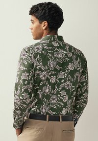 Vihreä kukkapaidassa on valkoisia kasvipainatuksia, pitkät hihat, kaulus ja slim fit -design. Täydentää beiget housut ja musta vyö.