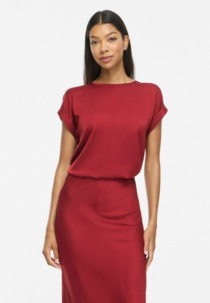 Un mannequin aux longs cheveux foncés se tient devant un fond blanc, vêtu d’un haut rouge à manches courtes et d’une jupe assortie arrivant aux genoux.