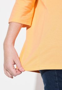 Kurzärmliges, gelbes T-Shirt aus weichem Stoff, mit einem leicht gewölbten Saum und sichtbaren Nähten, kombiniert mit dunkelblauen Jeans.