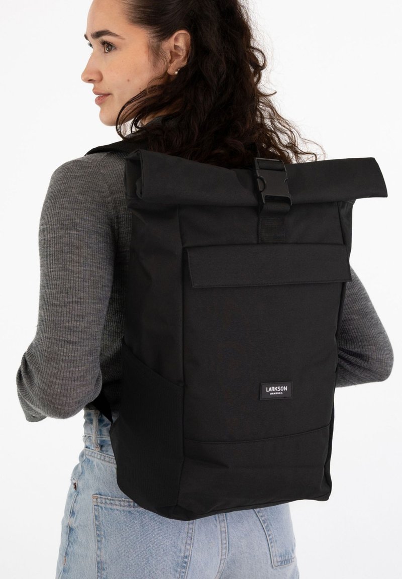 Jeune femme aux cheveux bouclés portant un pull gris et un jean bleu clair, portant un grand sac à dos noir à rabat étiqueté "Larkson."