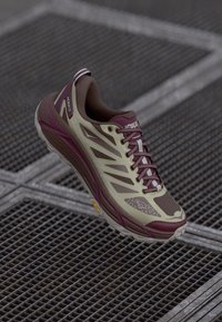 Scarpe da corsa verde chiaro e bordeaux con tomaia in rete, suola in gomma e un marchio HOKA ben visibile, caratterizzate da un design curvo e dettagli testurizzati.