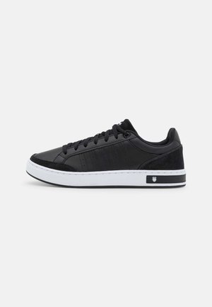 Zwarte lage sneaker met witte zool, veters aan de voorkant en subtiele strepen aan de zijkant, zijaanzicht op een effen lichte achtergrond.