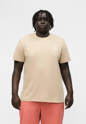 Άνδρας με σκούρο δέρμα και πλεξούδες που φοράει μπεζ μπλουζάκι Polo Ralph Lauren και κοραλί ροζ σορτς, σε λευκό απλό φόντο.