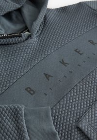 Grå hættetrøje med quiltet tekstur, glat stofpanel og præget "BAKER BY TED BAKER" logo på tværs af brystet.