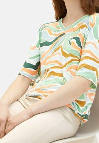 Femme portant un haut ample à manches courtes avec un motif abstrait multicolore, pantalon beige, assise les mains posées sur les genoux.
