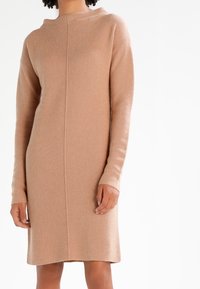 En modell med lockigt hår bär en lång, beige stickad klänning med hög hals och ribbad textur, stående mot en enfärgad vit bakgrund.