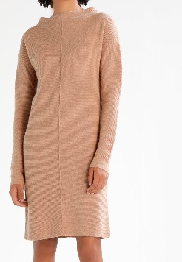 En modell med lockigt hår bär en lång, beige stickad klänning med hög hals och ribbad textur, stående mot en enfärgad vit bakgrund.