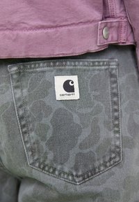Paarse spijkerjas met kraag bovenop grijze jeans met subtiele print, met een wit vierkant label met zwart logo op de achterzak.