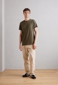 Oliven grønn t-skjorte med kort erme, beige løse bukser, svarte sneakers med hvite aksenter, står på en tregulv mot en nøytral bakgrunn.