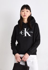 Juodas džemperis su dideliu baltu logotipu "Calvin Klein Jeans" priekyje. Turėtų kengūros tipo kišenę ir reguliuojamą raištelį ant gaubto.