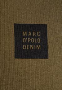 Olivegrüner Stoff mit einem schwarzen quadratischen Etikett, auf dem in großen gelben Buchstaben "MARC O'POLO DENIM" steht.