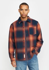 Camisa a cuadros en tonos azules, naranjas y marrones. Con botones en la parte delantera, cuello, un bolsillo en el pecho y mangas largas. Tejido texturizado.