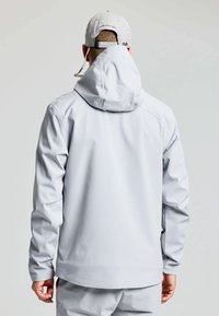 Giacca impermeabile grigia chiara con cappuccio, dotata di polsini elasticizzati, texture liscia e design comodo. La vista posteriore mostra dettagli minimi.
