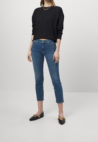 Sudadera negra corta, pantalones vaqueros ajustados azules de longitud ankle y zapatos planos negros con detalle de cadena dorada. Tejido suave y diseño ceñido.