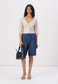 Top bege de gola em V com nervuras e mangas bufantes, combinado com culotes de jeans azul. A modelo segura uma bolsa preta texturizada, usando sandálias bege com tiras.