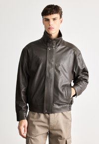 Pruunist nahast jakk, millel on kõrge krae, tõmblukud ja lõdva lõikega, ribakudumisega varrukad ja hem. Sile tekstuur, ilma mustriteta.