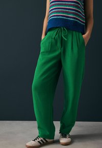 Groene, loszittende broek met een elastische tailleband en zijzakken, gecombineerd met een gestreepte trui in meerdere kleuren en beige sneakers.
