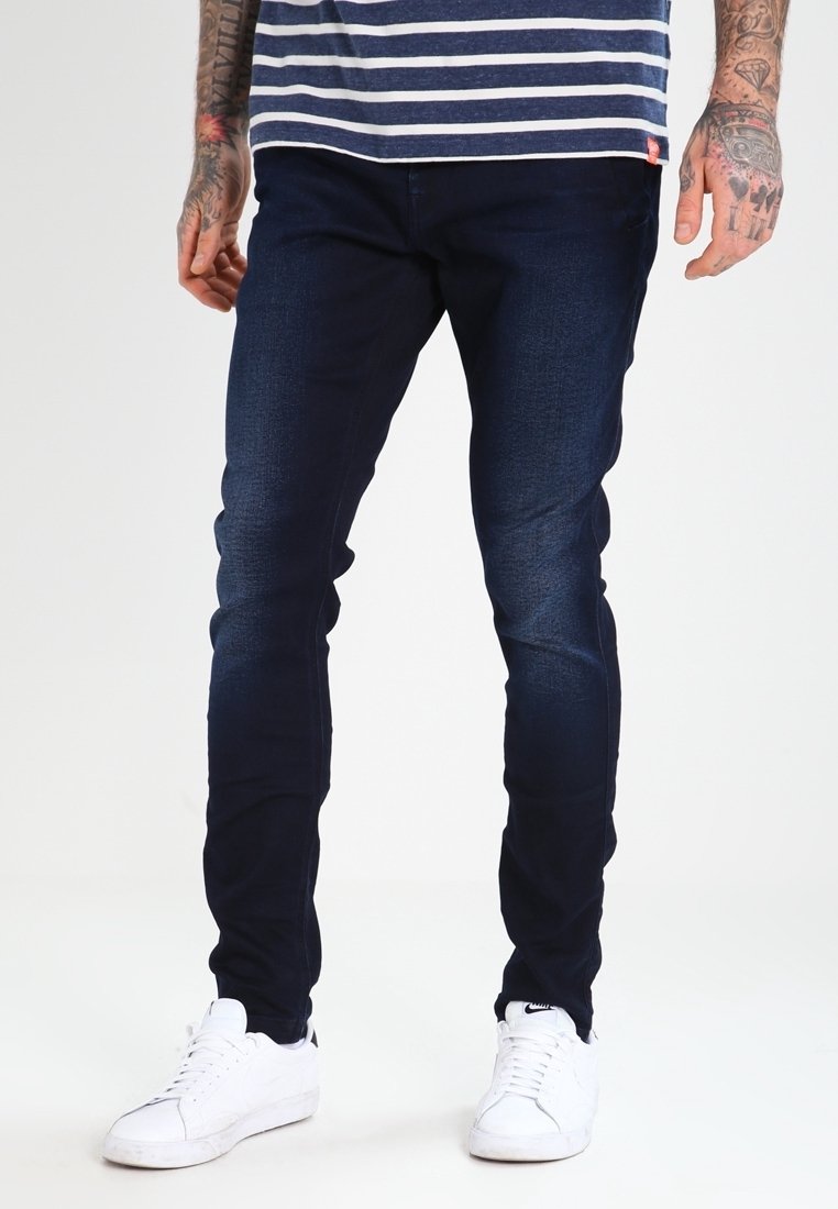 Man med mörkblå slim-fit jeans, vita sneakers och en randig marinblå och vit skjorta, med tatuerade underarmar synliga.