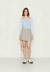 Camisola de mangas longas azul clara com detalhes franzidos, saia mini xadrez em bege e castanho, sandálias brancas de dedo e uma pequena mala rosa.