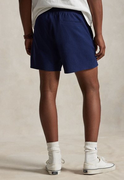 Marineblå shorts med elastisk midje, baklomme og glatt tekstur, kombinert med hvite høye sneakers og ribbestrikkede sokker.